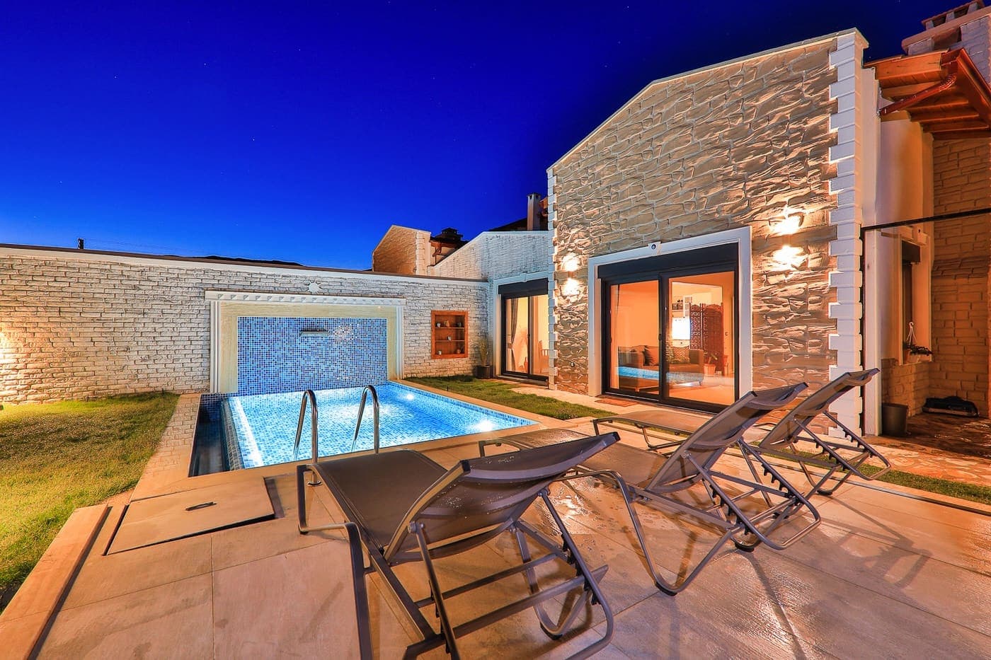 Akşam ışıklandırmalı havuz başı, şezlonglar ve taş cepheli villa.