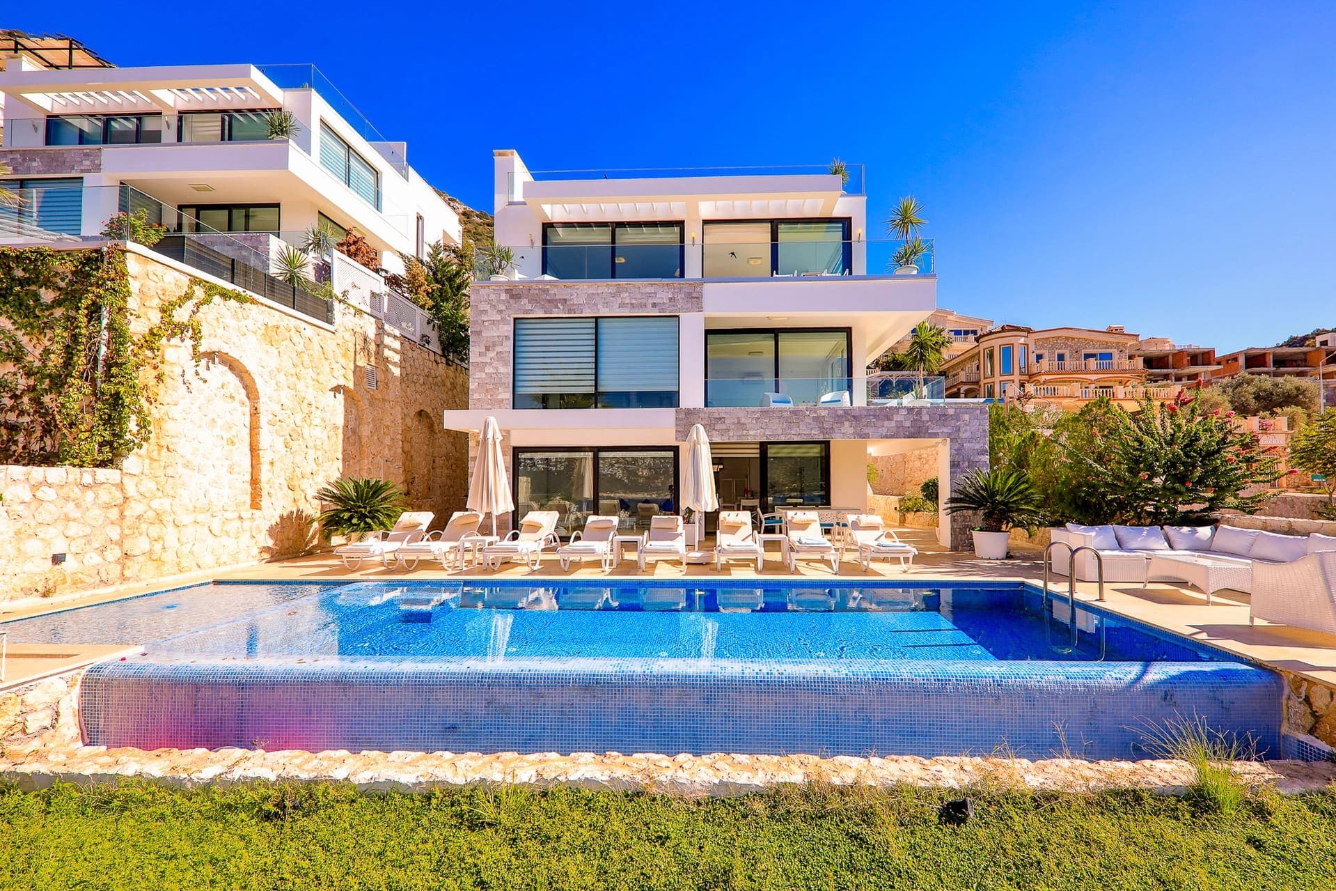 Çok katlı modern villanın önünde şezlonglu yüzme havuzu ve oturma alanı.