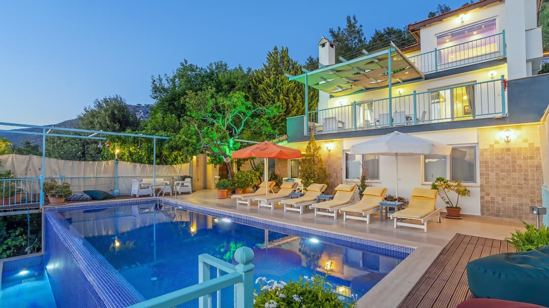 Yeşillikler arasında, havuz başında şezlonglar ve turuncu güneş şemsiyeleri olan iki katlı villa.