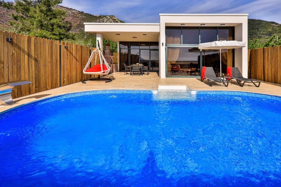Önünde mavi havuz ve şezlonglar olan, ahşap çitlerle çevrili tek katlı modern villa.