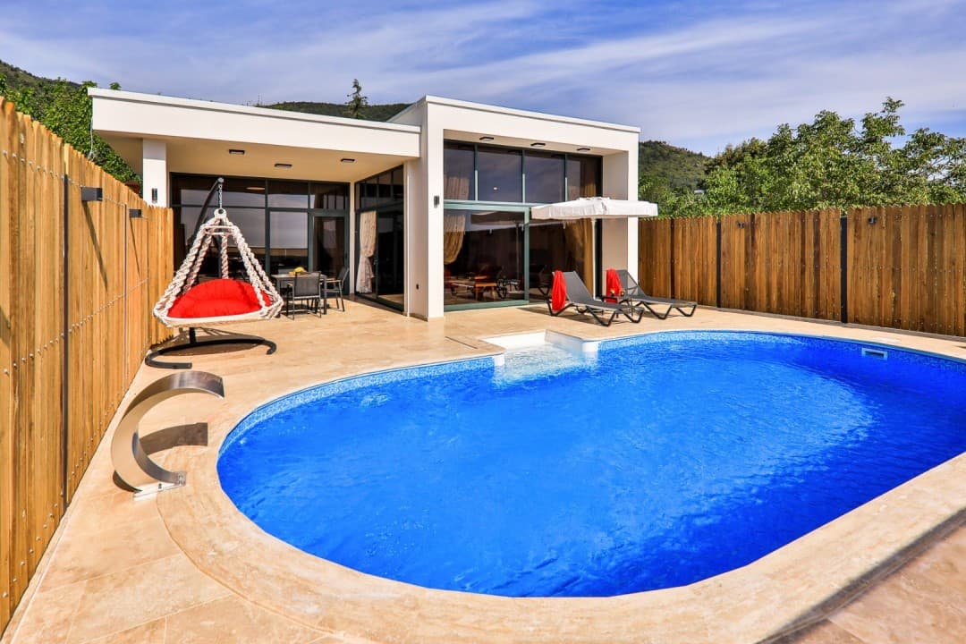 Önünde havuzu ve ahşap çitlerle çevrili terasında kırmızı minderli salıncak bulunan modern villa.