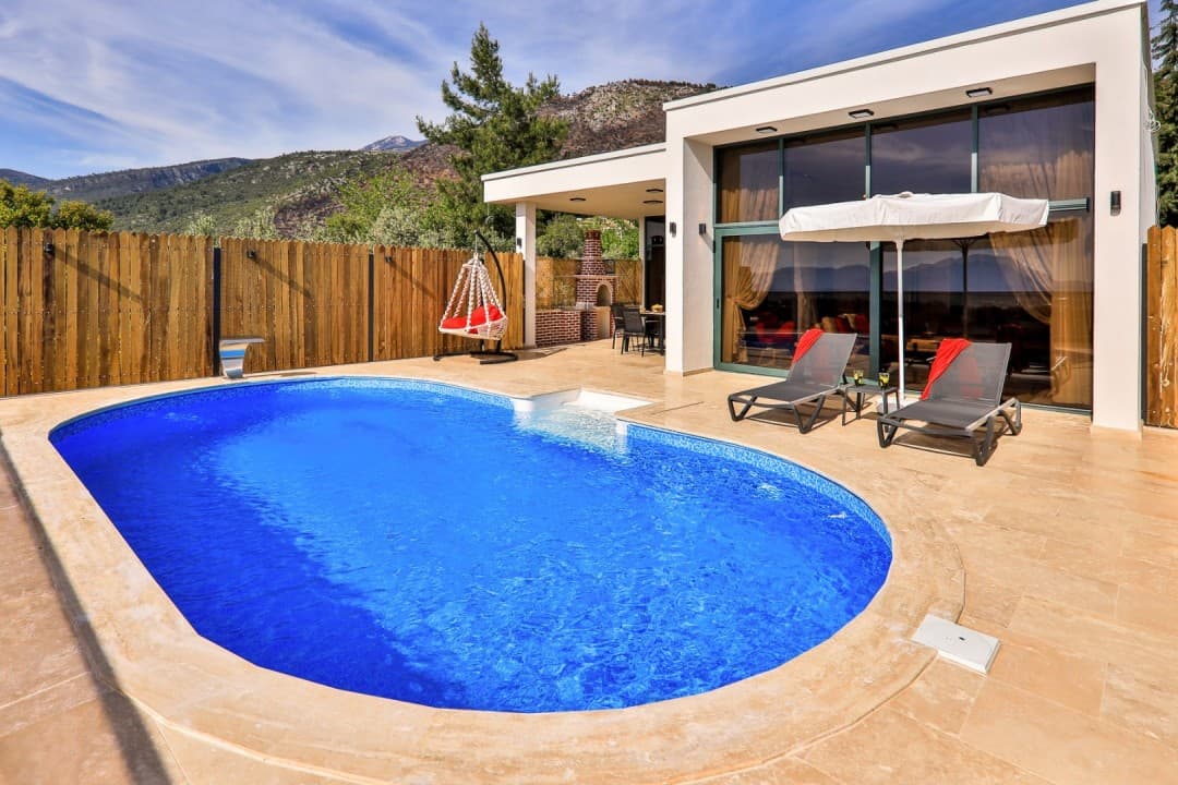 Ahşap çitlerle çevrili bahçede oval havuz ve şezlonglar bulunan modern villa.