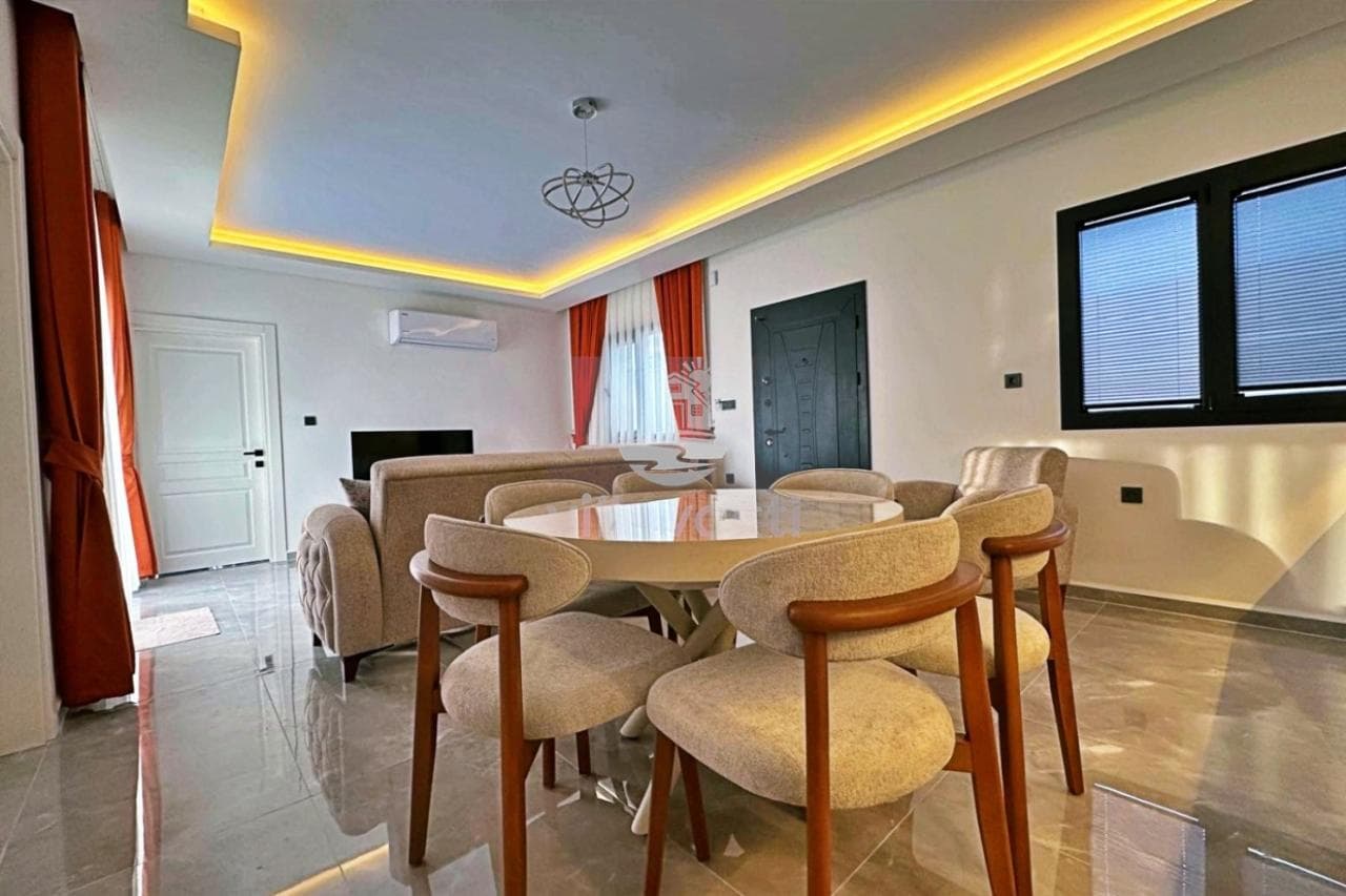 Aydınlatmalı tavanı olan geniş salonda yemek masası ve sandalyeler.