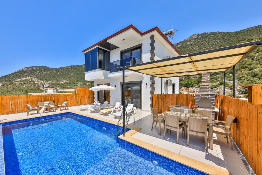 Özel havuzlu, bahçesinde yemek masası, barbekü ve şezlonglar bulunan beyaz dış cepheli iki katlı villa.