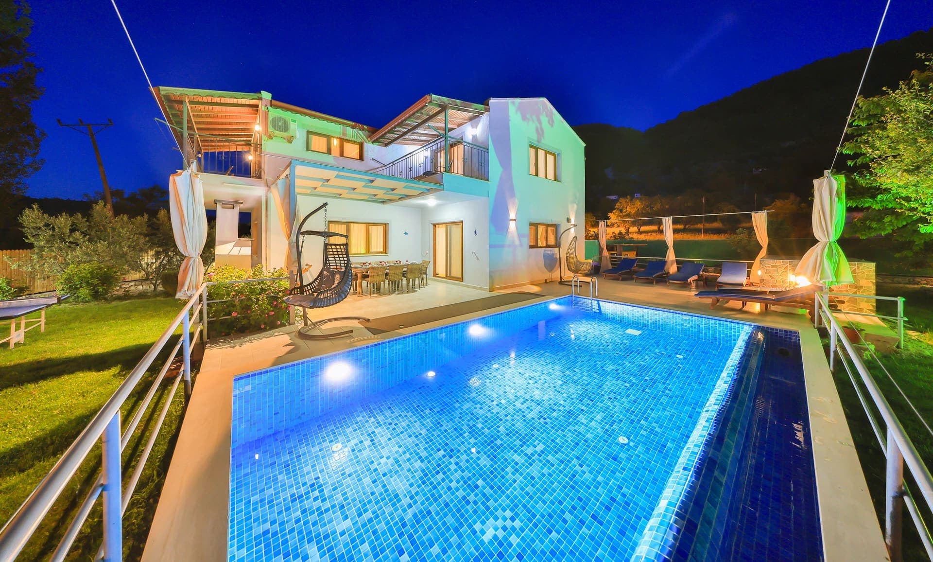 Akşam aydınlatmalı havuz ve şezlongları olan iki katlı villa.