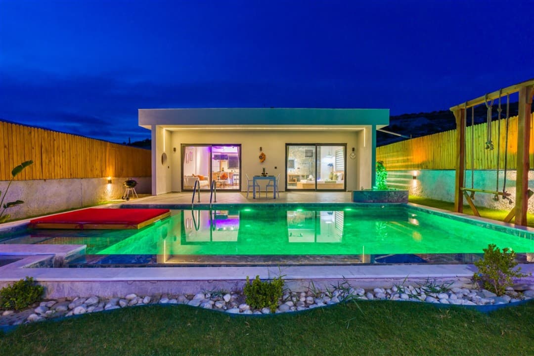 Gece vakti yeşil ışıklandırmalı havuzu ve önünde ahşap salıncak bulunan müstakil villa.