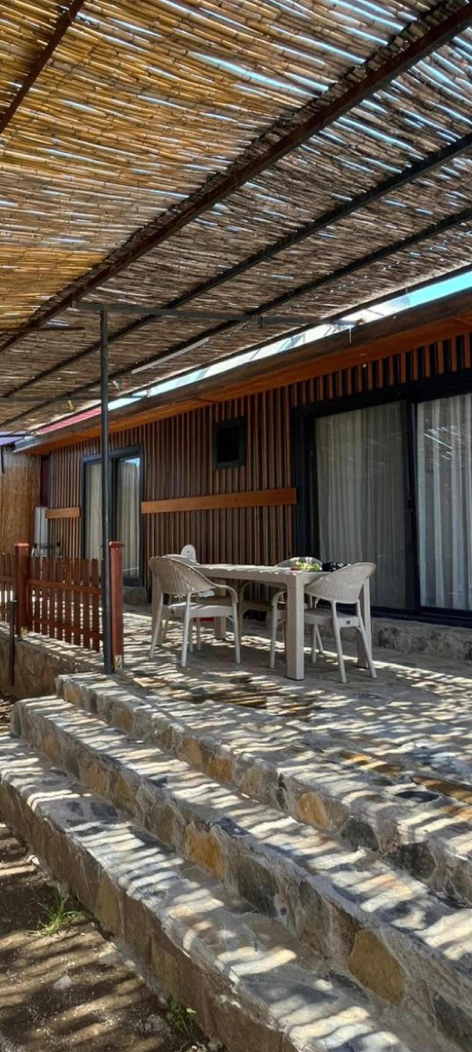 Ahşap çıtalı cepheye sahip villanın taş zeminli verandasında masa ve sandalyeler var.