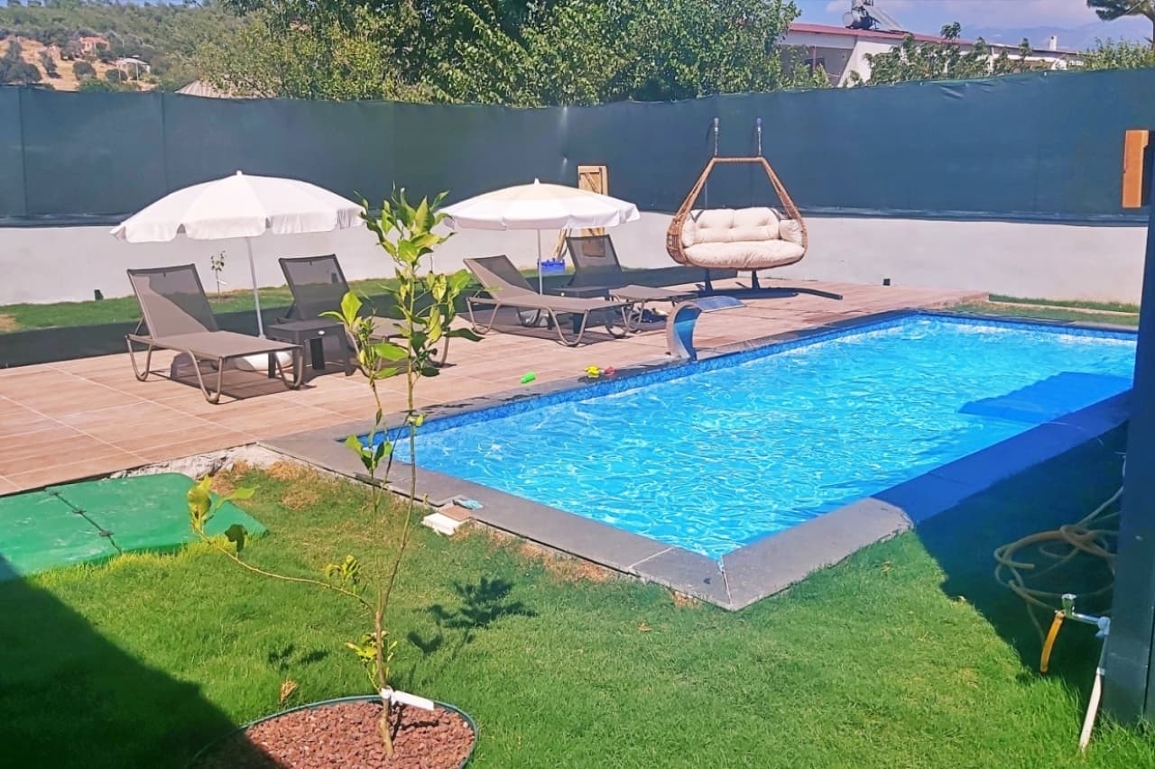 Ön planda küçük bir ağaç, arkada mavi yüzme havuzu ve şezlonglu teras.