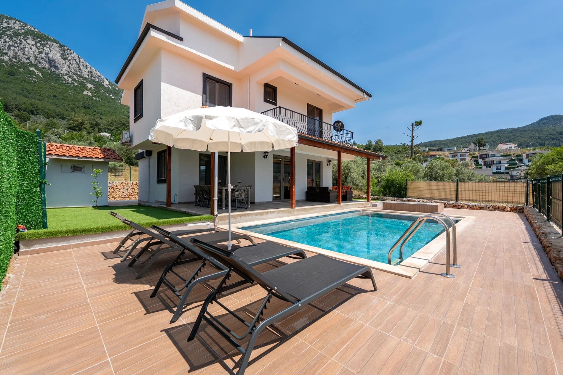 Havuz kenarında şezlonglar, beyaz şemsiye ve arkada dağ manzaralı villa dış cephesi.