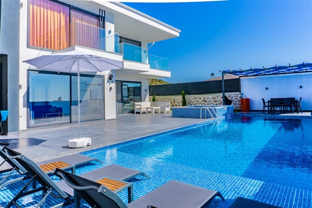 Güneş şemsiyesi ve şezlongların yer aldığı açık havuz ile beyaz katlı villa.