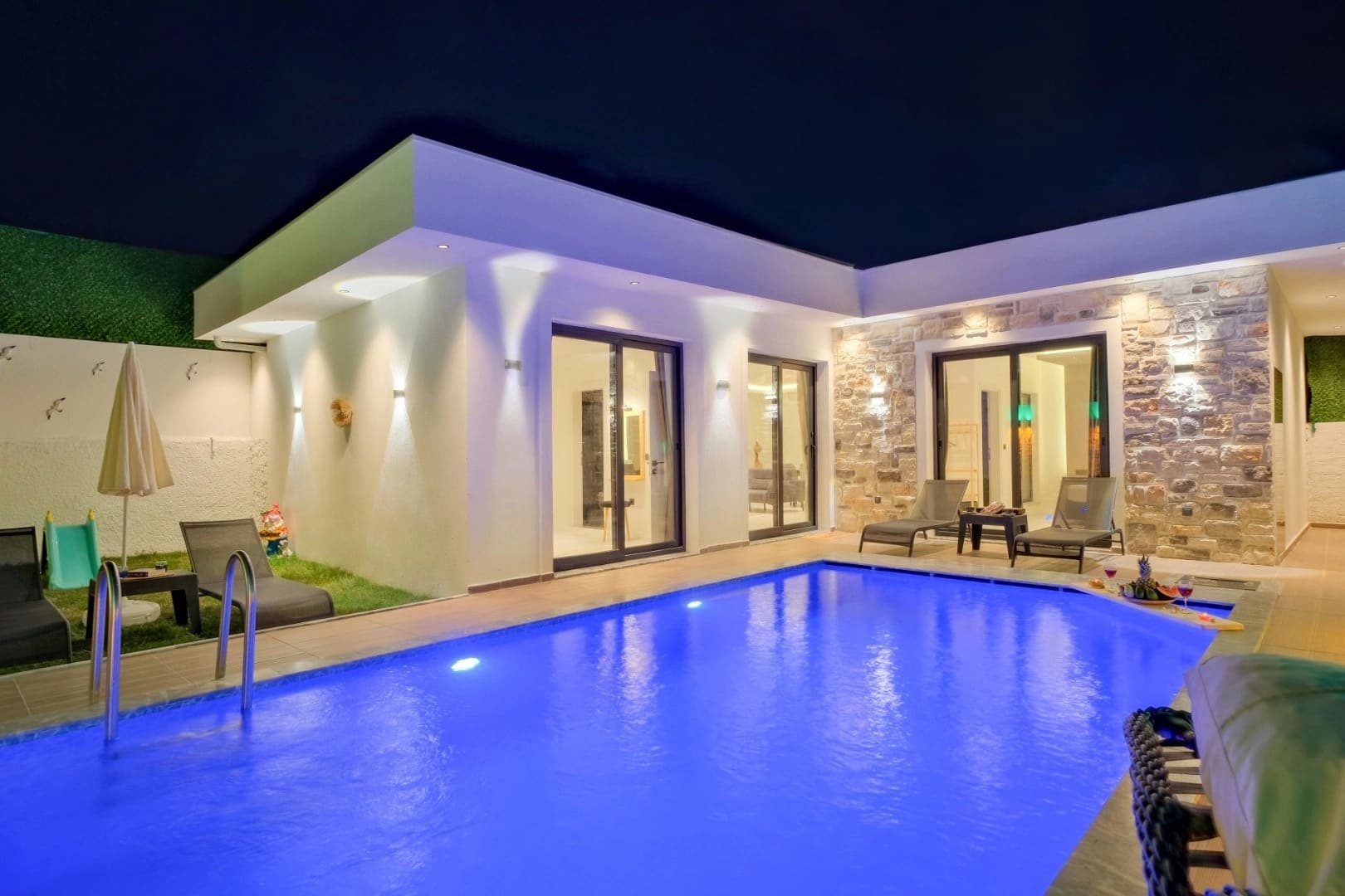 Aydınlatmalı havuzun başında şezlonglar bulunan, gece çekilmiş villa dış cephesi.