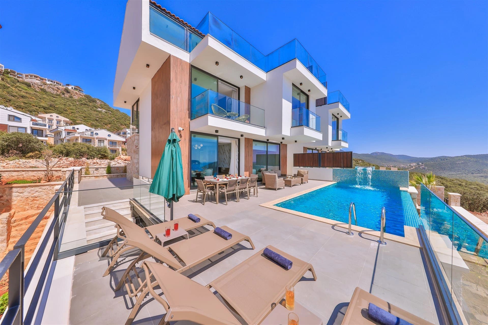 Geniş havuzlu terasta şezlonglar ve yemek masası bulunan iki katlı villa.