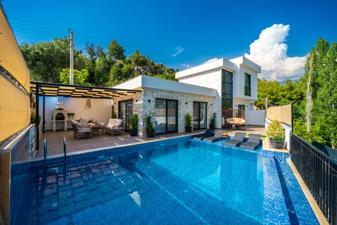 Önünde güneşlenme şezlongları ve gölgelikli oturma alanı bulunan, taş kaplamalı modern özel havuzlu villa.