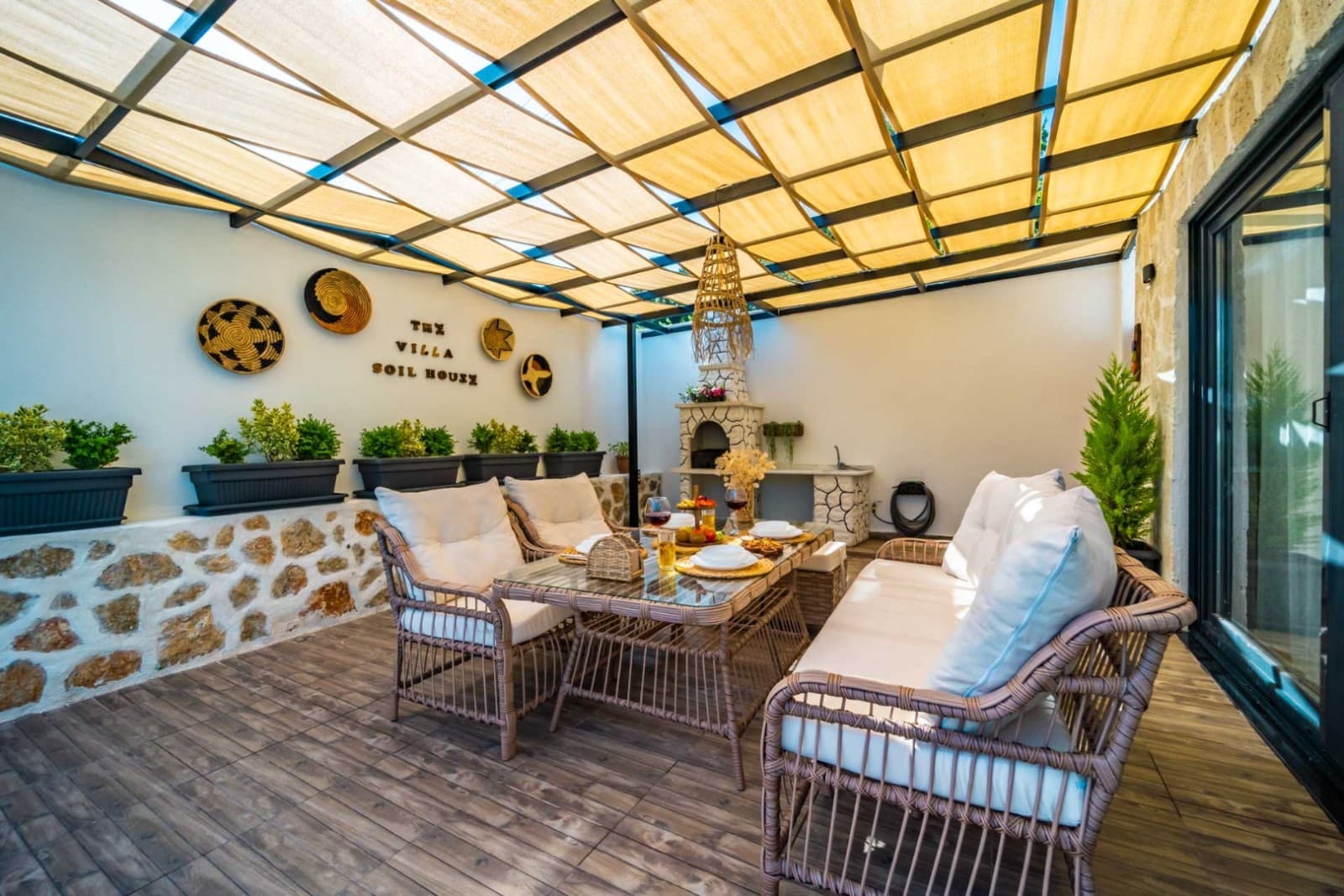 Ahşap pergola gölgelikli, hasır oturma grubu ve yemek masası olan taş duvarlı teras.