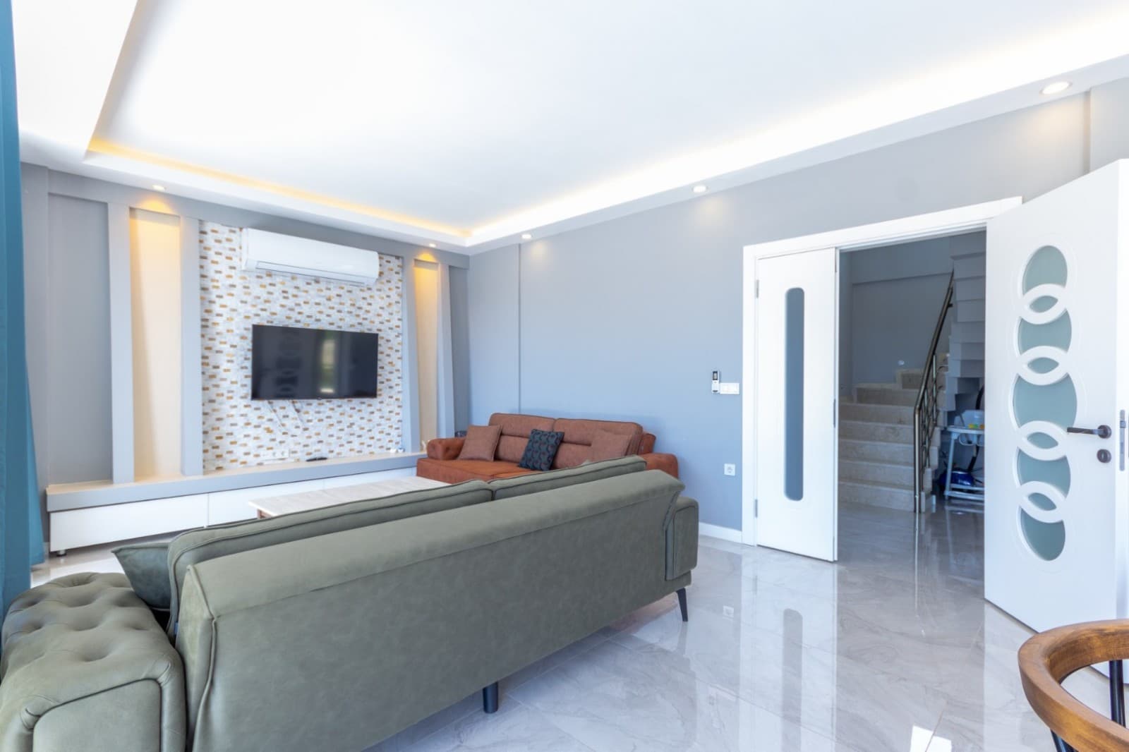 Gri duvarlı salonda yeşil kanepe, kahverengi koltuk ve TV ünitesi.