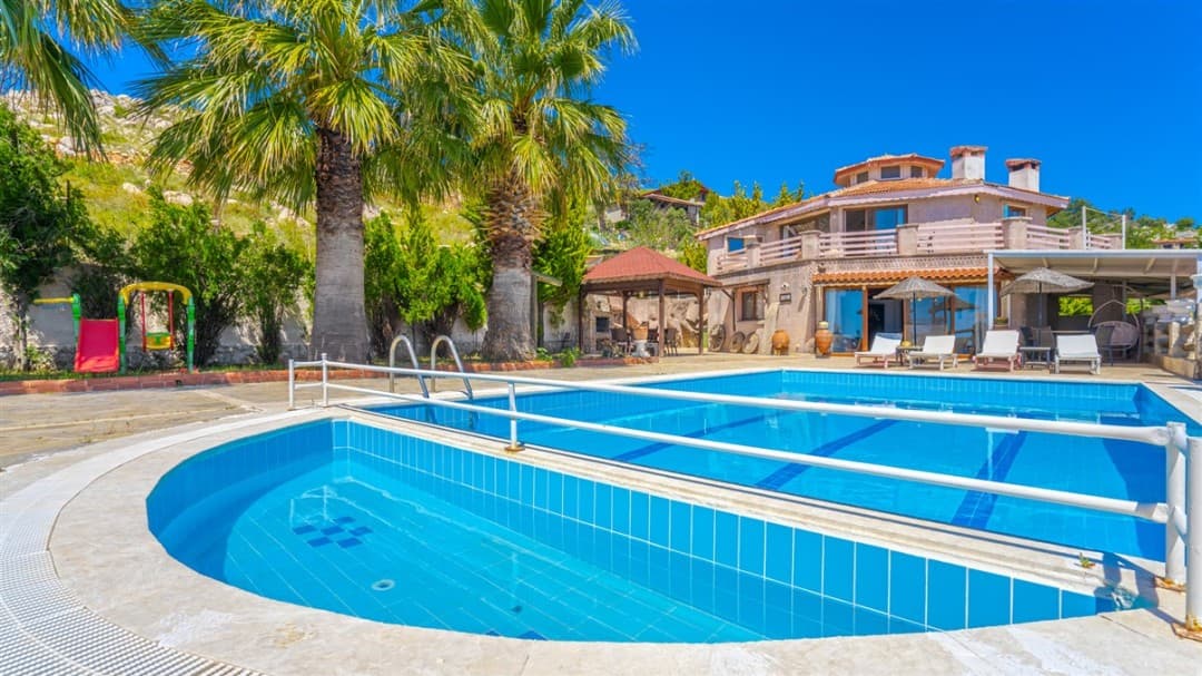 Bahçesinde palmiye ağaçları, çocuk parkı ve şezlonglar bulunan, özel havuzlu iki katlı taş villa.