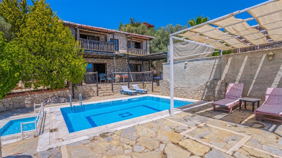 Taş kaplama iki katlı villa, önünde yüzme havuzu, şezlonglar ve gölgelikli oturma alanı.
