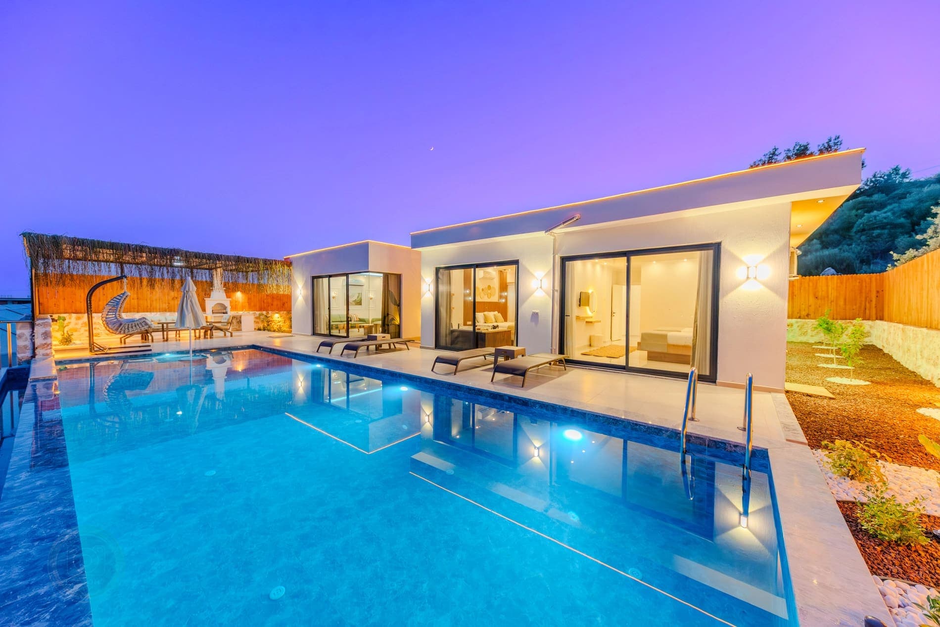 Akşam ışıklandırmalı havuzun başında şezlonglar ve tek katlı villa yer alıyor.