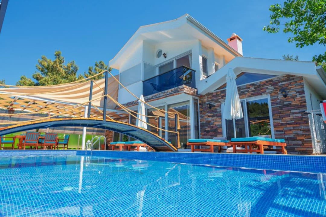 Taş kaplamalı iki katlı villa, önünde mavi mozaik havuz, ahşap köprü ve güneş şemsiyeli şezlonglar bulunuyor.