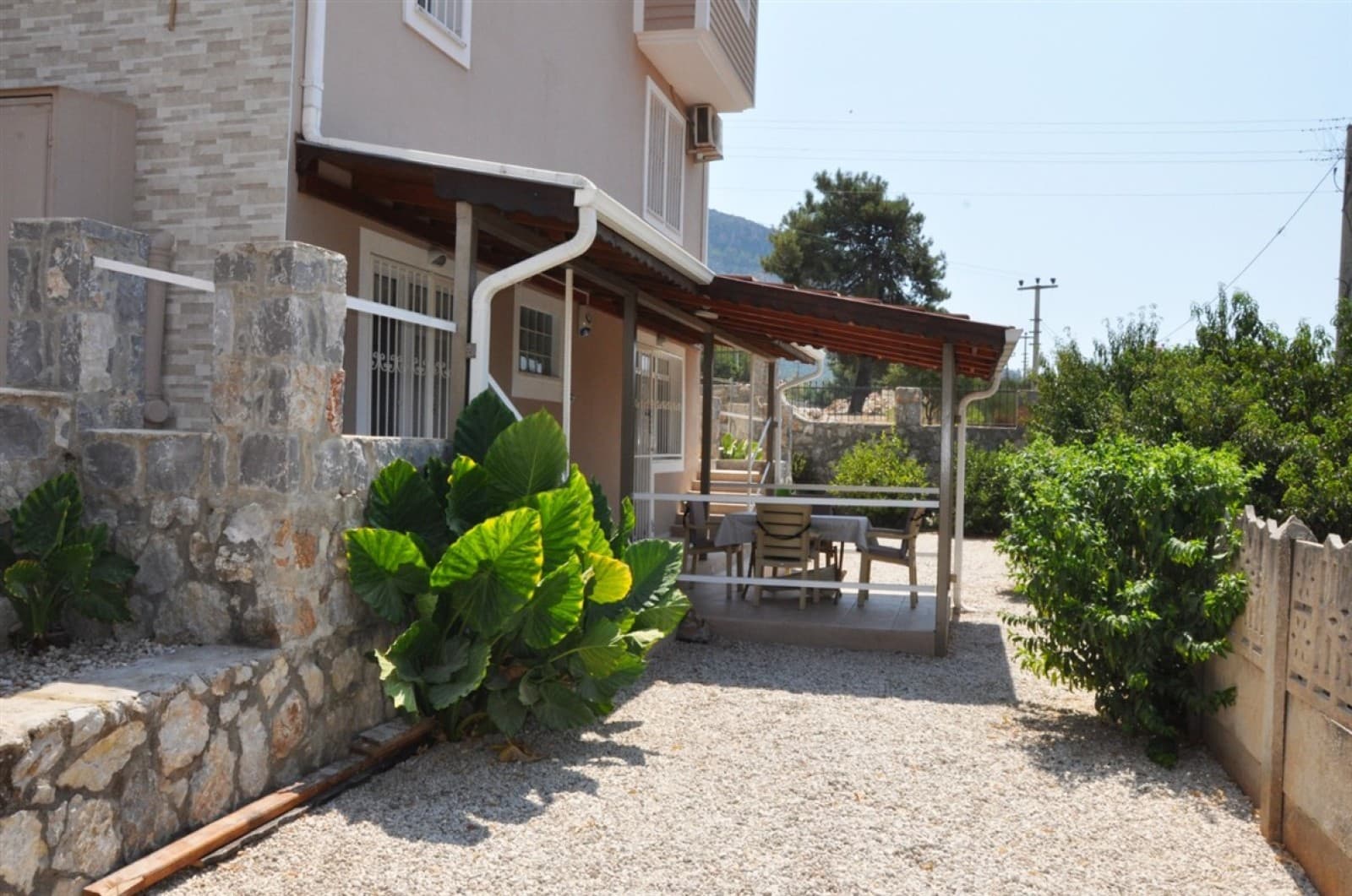 Çakıl taşlı bahçede üzeri kapalı teras alanı, oturma grubu ve yeşil bitkiler bulunan villa dış cephesi.