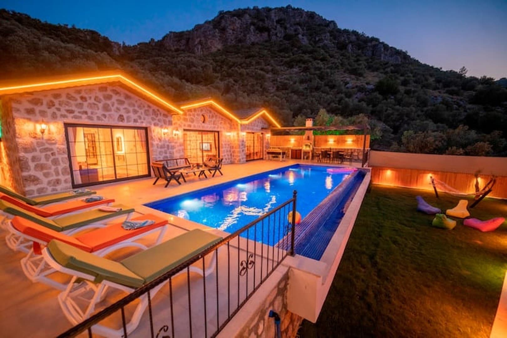 Akşam saatlerinde aydınlatılmış taş villa, önünde yüzme havuzu, şezlonglar ve çim bahçesi bulunuyor.