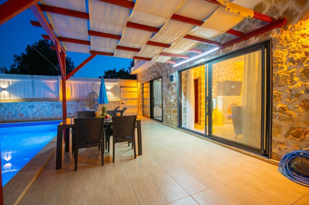 Akşam vakti aydınlatmalı havuz kenarında, gölgelikli terasta bulunan yemek masası ve taş duvarlı villa.