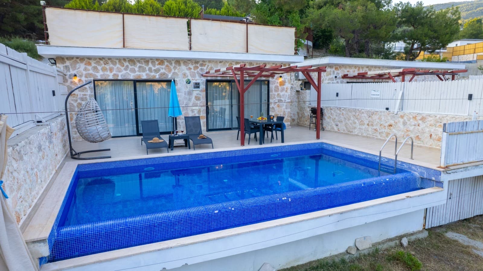 Taş cepheli villanın önünde mavi havuz, şezlonglar ve veranda alanında masa takımı.