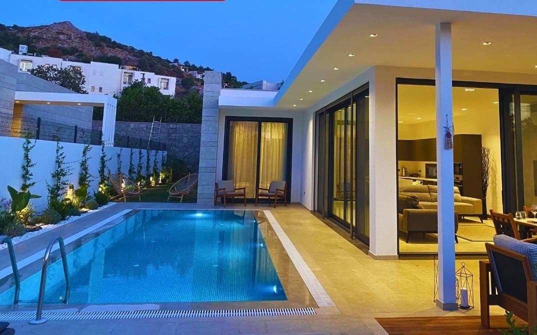 Aydınlatmalı havuz kenarında oturma grubu ve modern villanın cam cephesi görülüyor.