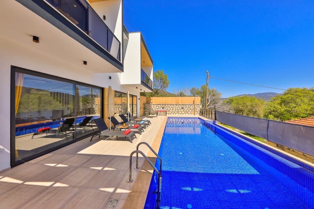 Açık havada masmavi uzun bir havuz ve kenarında şezlonglar bulunan modern villa dış cephesi.