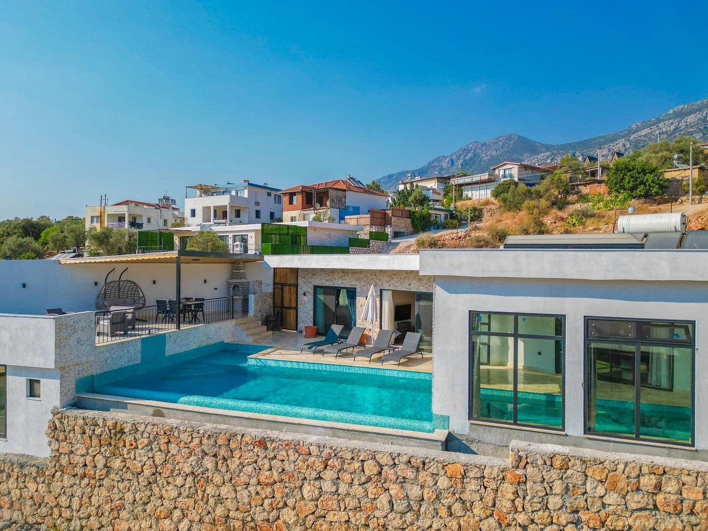 Dağ ve yerleşim yeri manzaralı, önünde taş duvar ve havuz bulunan modern villa dış cephesi.