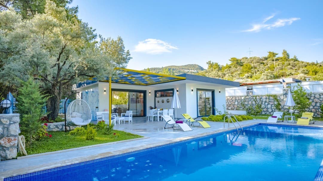 Önünde havuz bulunan, sarı gölgelikli verandasında oturma grubu ve şezlonglar yer alan beyaz tek katlı villa.