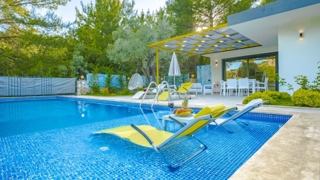 Havuz içinde sarı şezlonglar, kenarda masa, arkada beyaz villa ve ağaçlar yer alıyor.