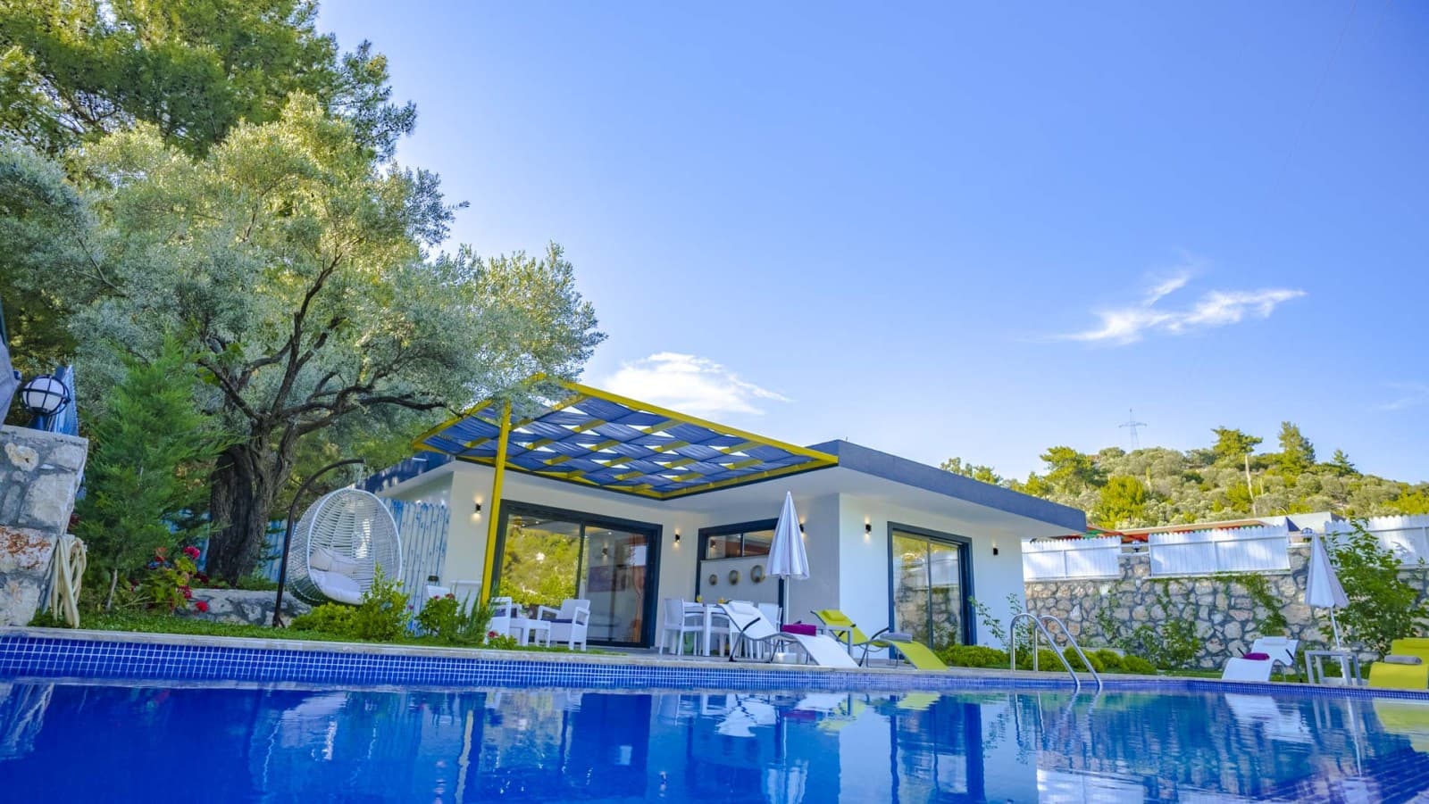 Mavi yüzme havuzu ve önünde şezlonglar bulunan beyaz cepheli tek katlı villa.
