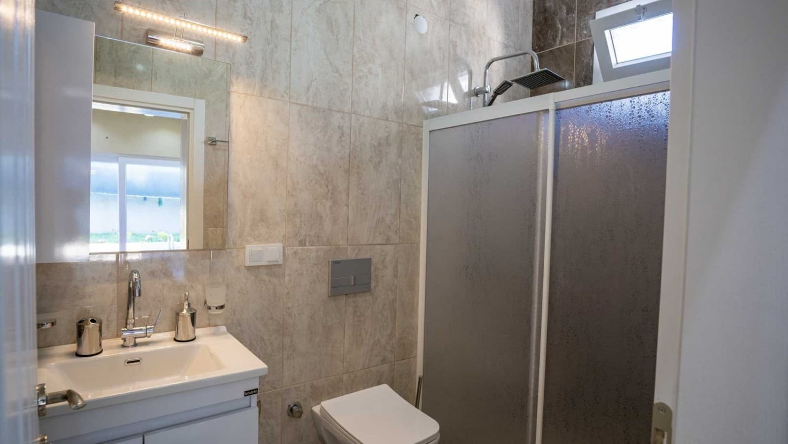 Bej tonlarında fayans kaplı, duşakabinli ve aynalı lavabosu olan modern banyo.