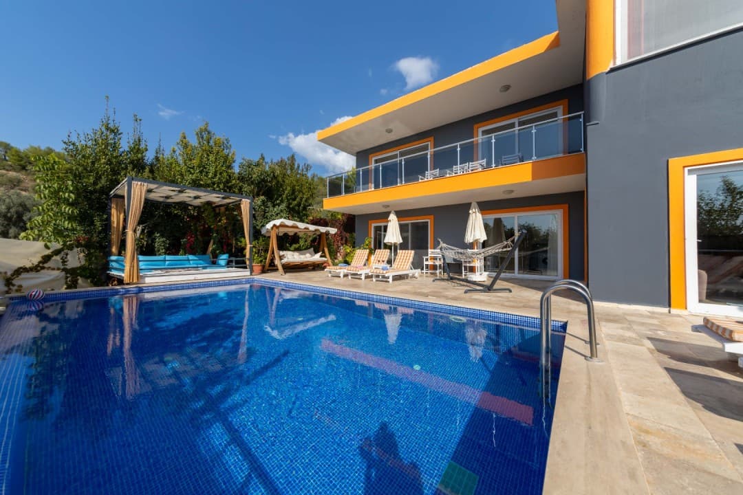 İki katlı gri ve sarı detaylı villanın önünde havuz ve şezlonglar.