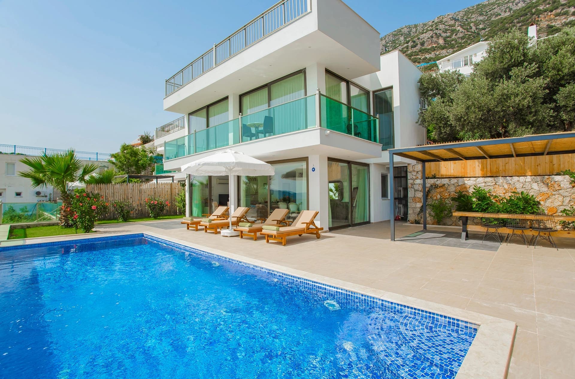 Önünde şezlonglar bulunan havuzlu modern beyaz villa ve yan tarafında gölgelikli oturma alanı.