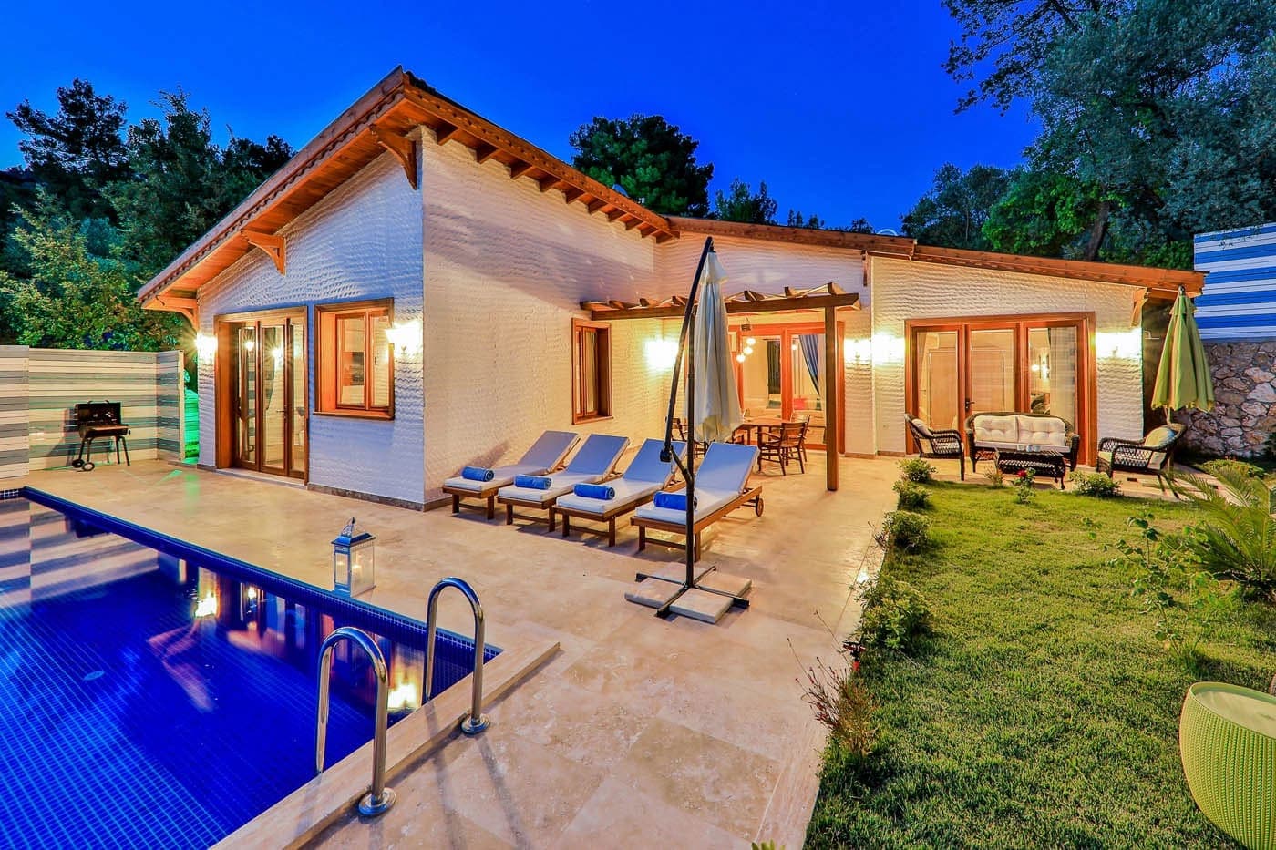 Gece aydınlatmalı, havuzlu ve şezlonglu tek katlı villa bahçesi.