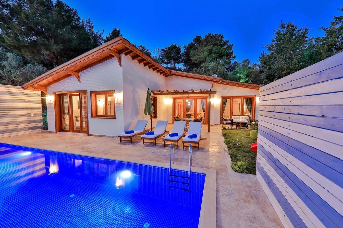 Gece aydınlatmalı, havuz kenarında şezlonglar bulunan tek katlı villa ve bahçesi.