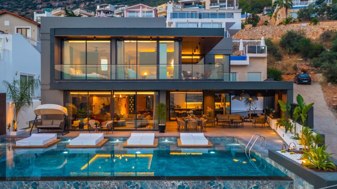Akşam ışıklarıyla aydınlatılmış, önünde havuz ve şezlonglar bulunan iki katlı modern villa.