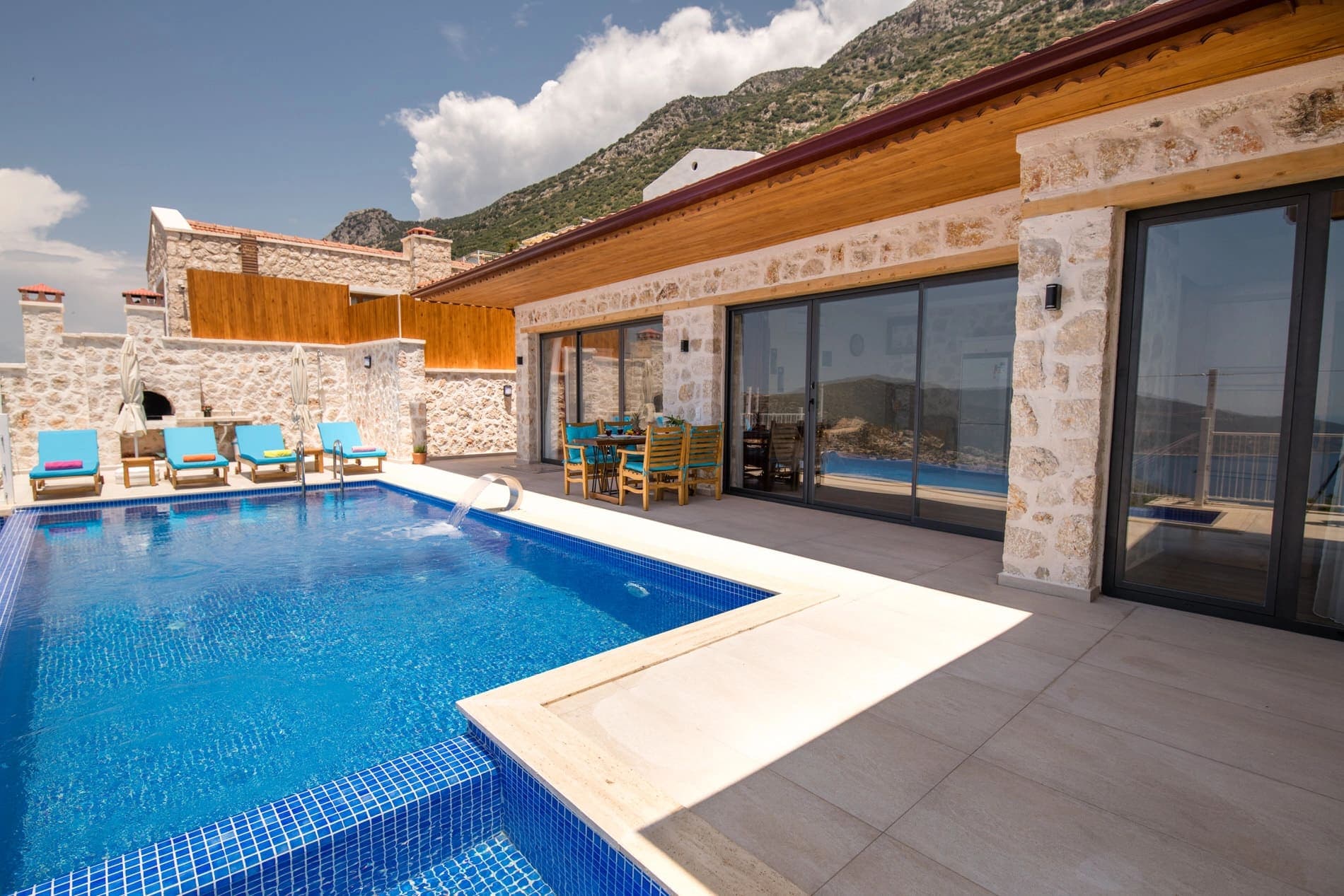 Taş duvarlı villa terasında mavi havuz, şezlonglar ve yemek masası.