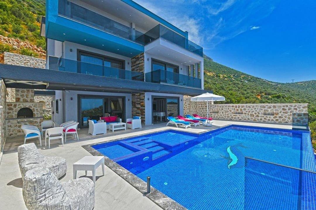 Mavi havuzlu ve balkonlu modern villanın önünde şezlonglar ve oturma grubu bulunuyor.