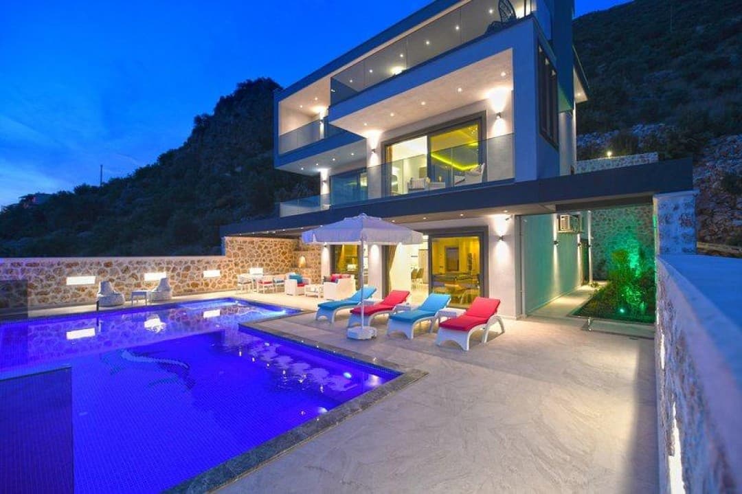 Akşam saatlerinde aydınlatılmış, havuzlu ve şezlonglu modern villanın dış cephesi.