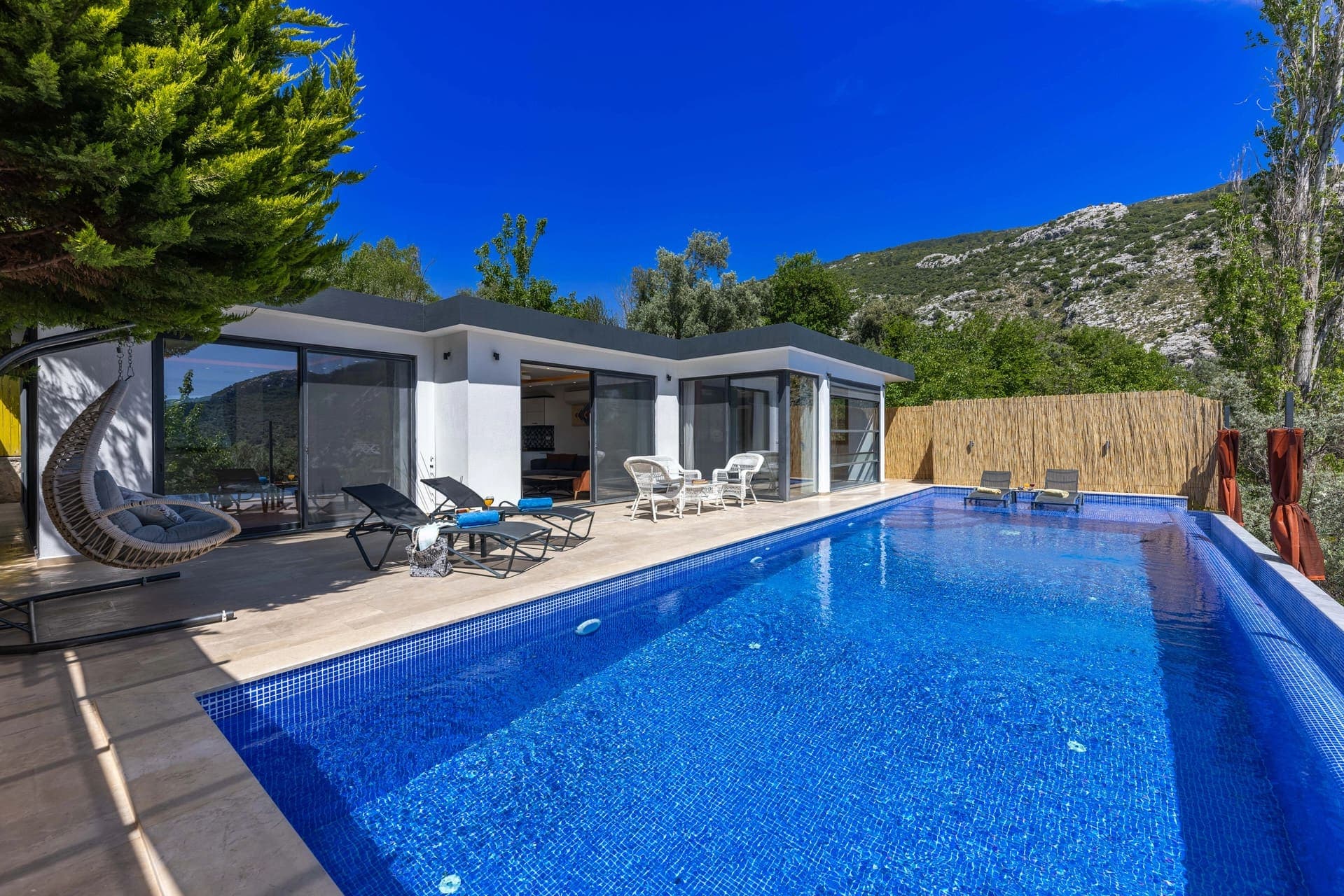 Mavi yüzme havuzu, şezlonglar, sallanan koltuk ve arkada dağ manzarası olan modern villa.