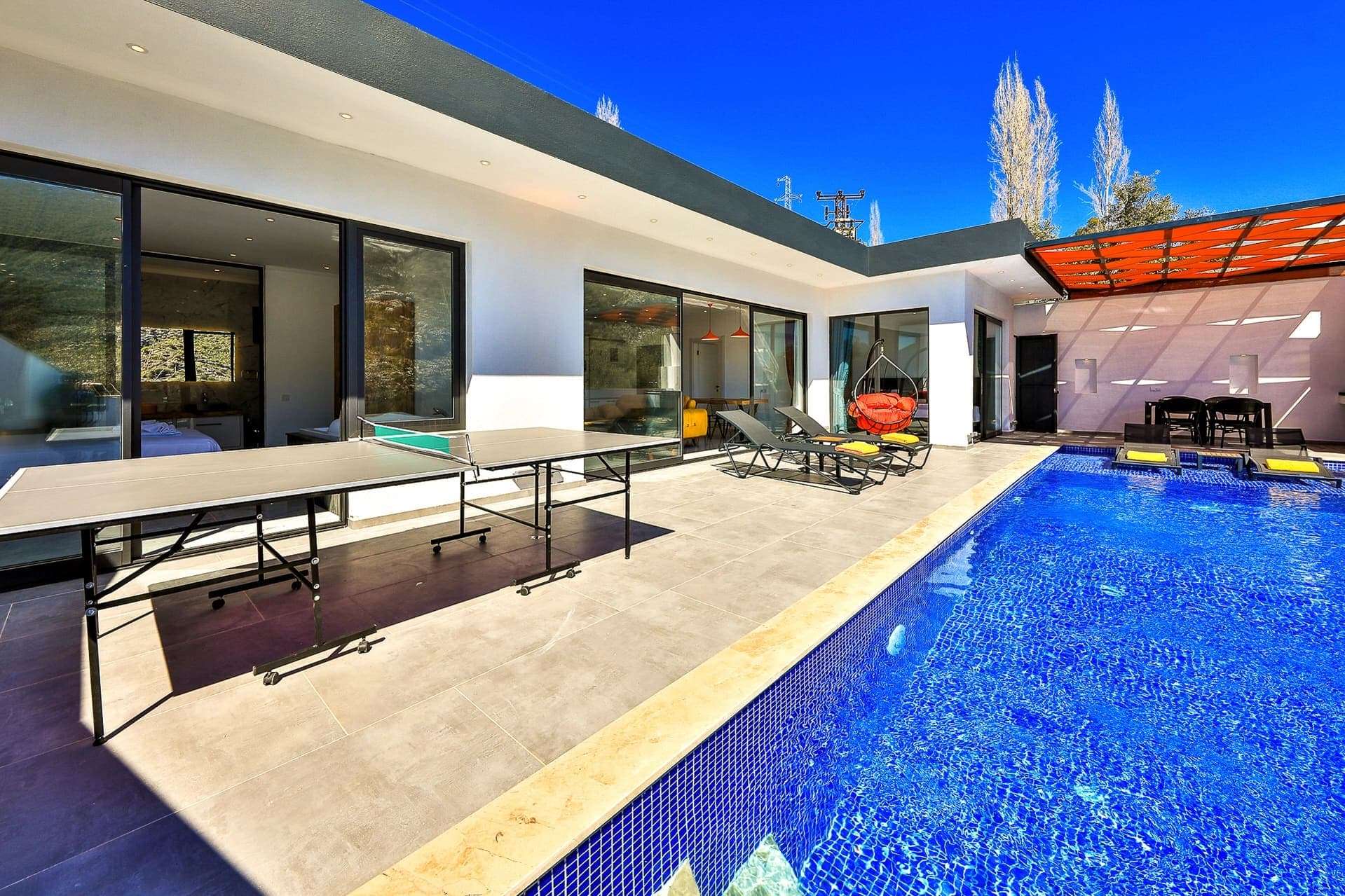 Havuz kenarında masa tenisi, şezlonglar ve oturma alanı bulunan modern villa bahçesi.