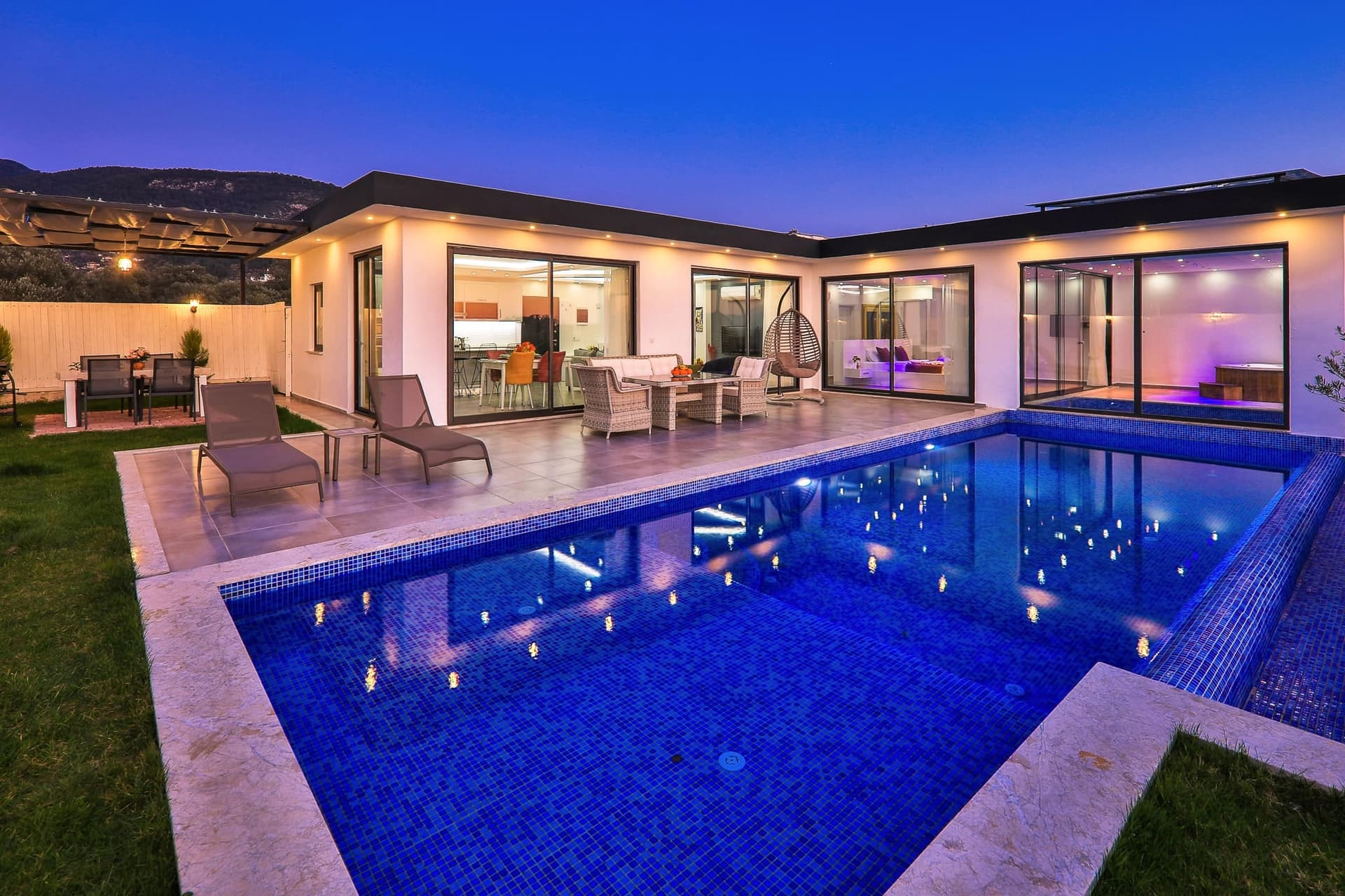 Gece aydınlatmalı, havuz başında oturma grubu ve şezlonglar bulunan modern villa bahçesi.