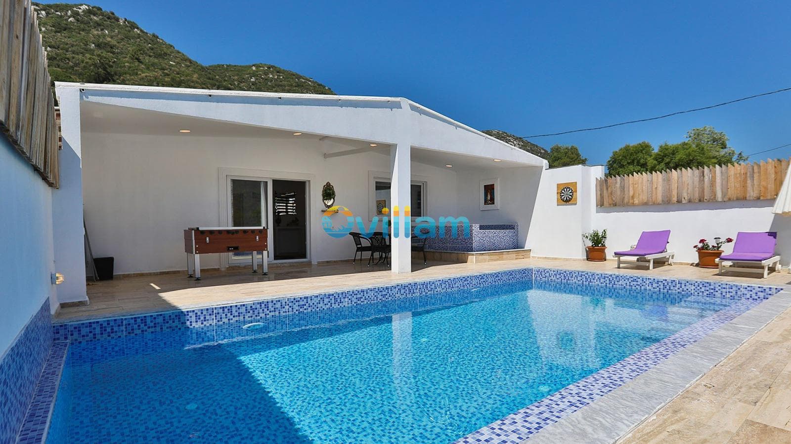 Mavi mozaik kaplı havuz, yanında mor şezlonglar ve masa tenisi bulunan villa bahçesi.