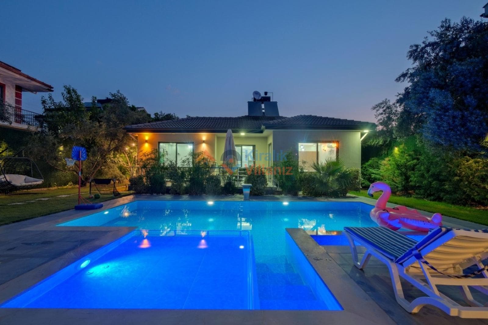 Gece aydınlatmalı havuz, flamingo deniz yatağı ve şezlongların olduğu villa bahçesi.