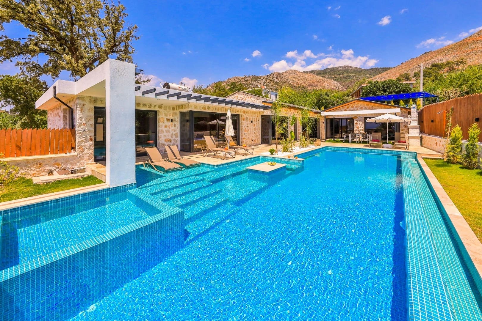 Taş mimarili, geniş havuzlu ve şezlonglu villa dış cephesi.