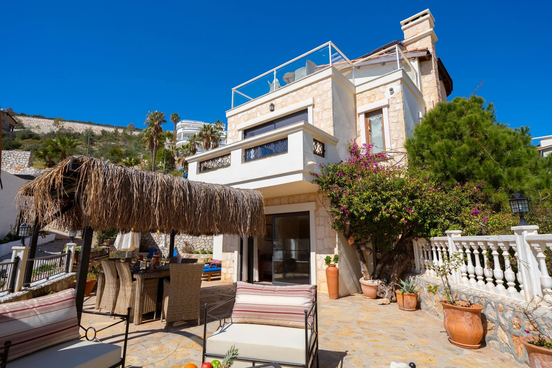 Hasır gölgelikli oturma alanı, çiçekli ağaçlar ve taş detaylı dış cepheye sahip villa.