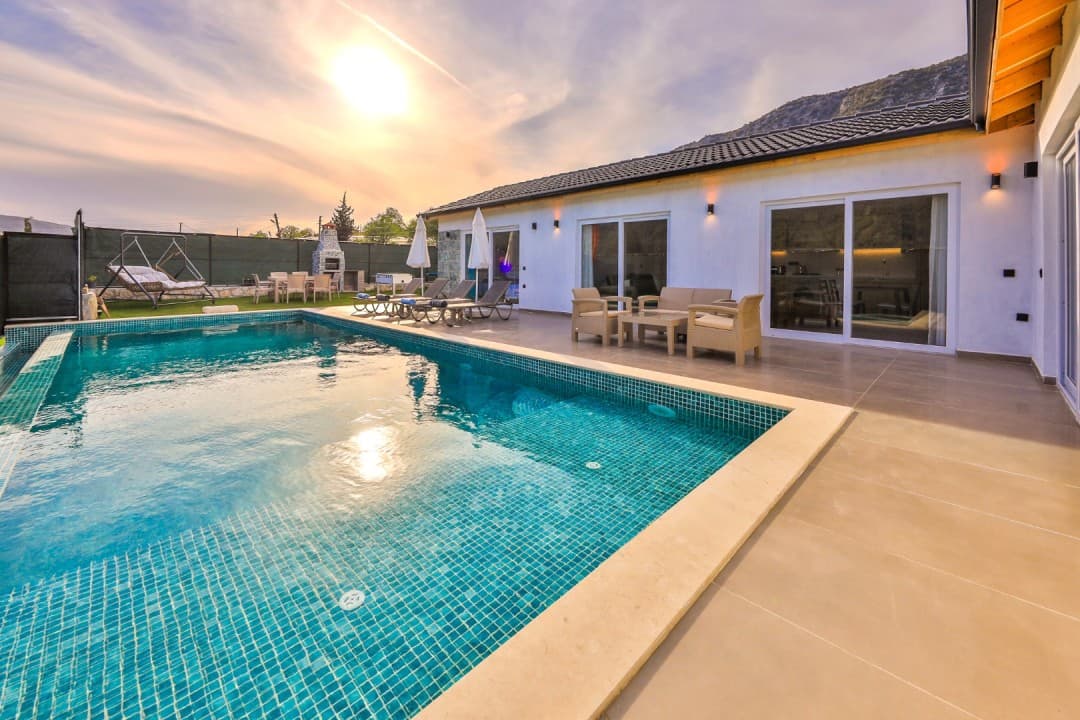 Gün batımında havuz başı oturma grubu ve şezlongları bulunan tek katlı beyaz villa.